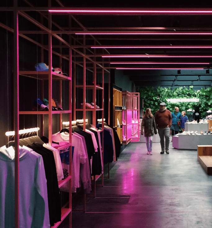 Modern retail store interior with stylish clothing displays, vibrant pink lighting, and shoppers exploring the space, emphasizing a contemporary shopping experience.