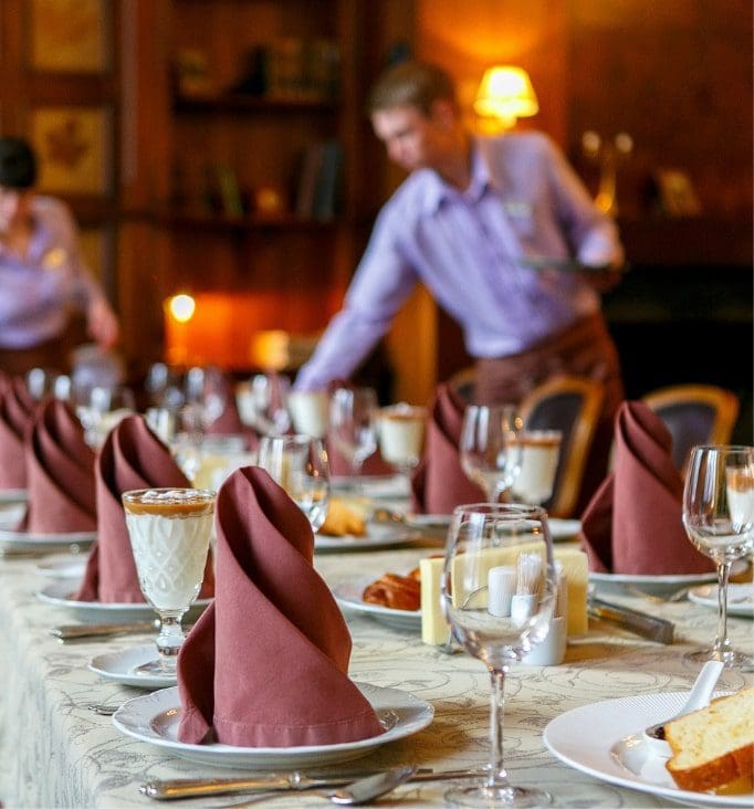 Waiters serving elegantly set dining table with folded napkins, glassware, and desserts in a hospitality setting.