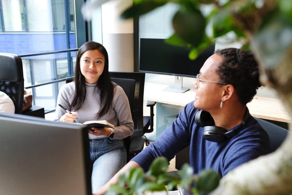 Two colleagues engaged in a discussion in a modern office setting, with one taking notes and the other wearing headphones, reflecting a collaborative and productive work environment focused on sustainability and responsible practices.