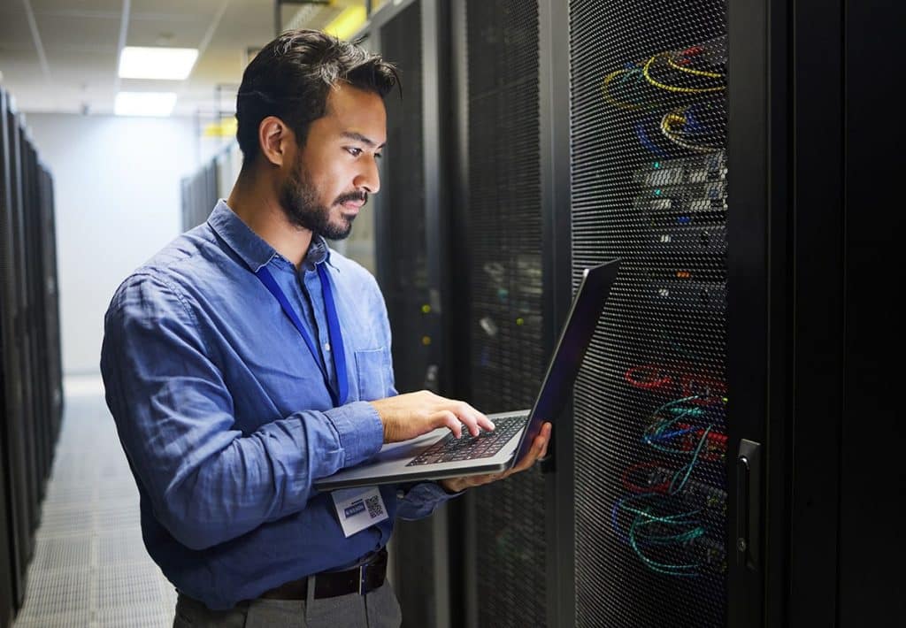 Man working on a laptop in a data center, managing server configurations and backup processes for data security and disaster recovery solutions.