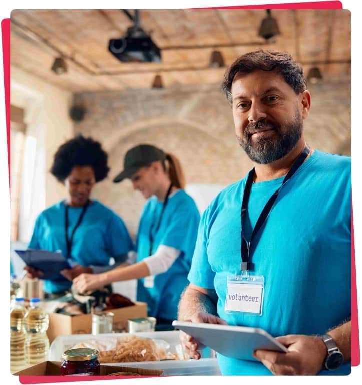 Volunteer in blue shirt smiling at camera, assisting in charity work with team members organizing supplies in background, emphasizing community support and teamwork in non-profit sector.
