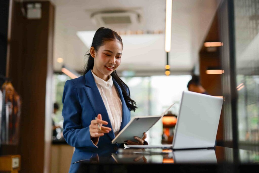 Asian businesswoman in a blue suit working on a laptop and tablet in a modern hospitality setting, reflecting Reliable Networks' IT support for the hospitality industry.