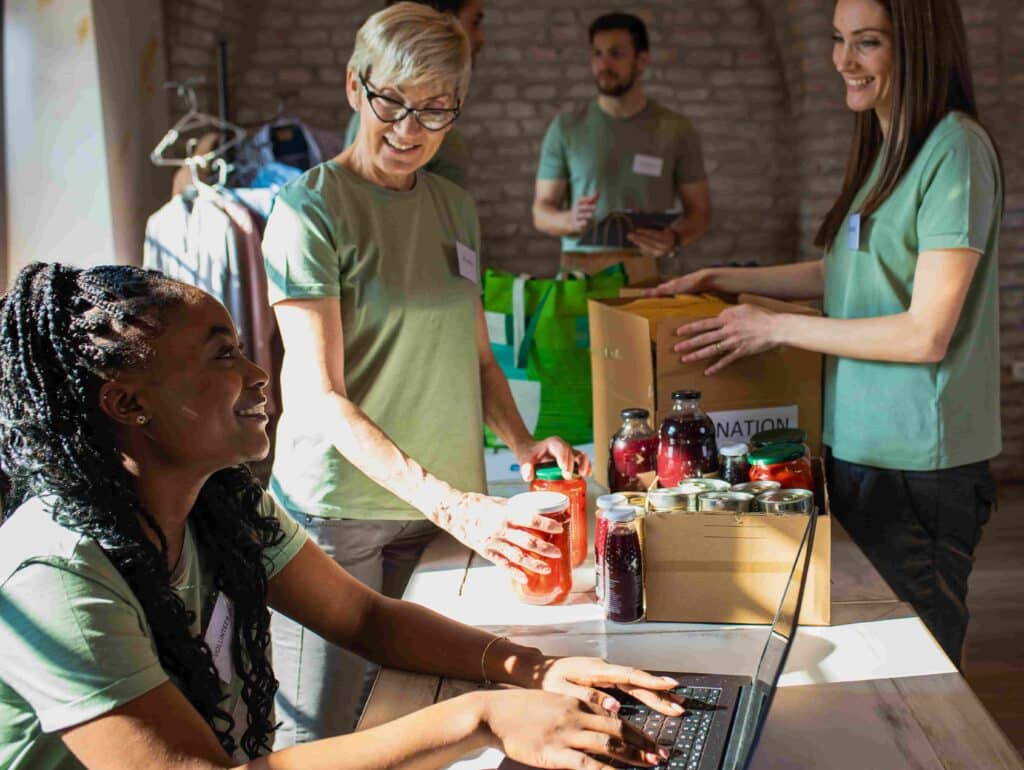 Group of volunteers in green shirts working together in a community charity setting, sorting donations and using a laptop for organization.