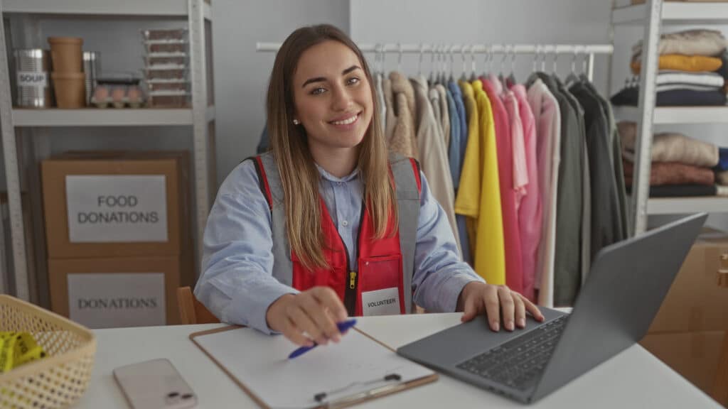 Volunteer smiling at a desk with a laptop, clipboard, and food donation boxes in a not-for-profit organization setting, emphasizing community support and digital transformation.