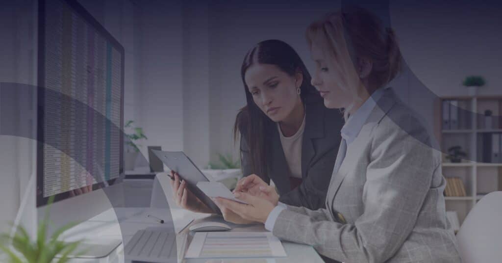 Two businesswomen discussing data on a tablet in a modern office, with a computer displaying financial information in the background, highlighting the importance of managed IT services for finance companies' security and compliance.